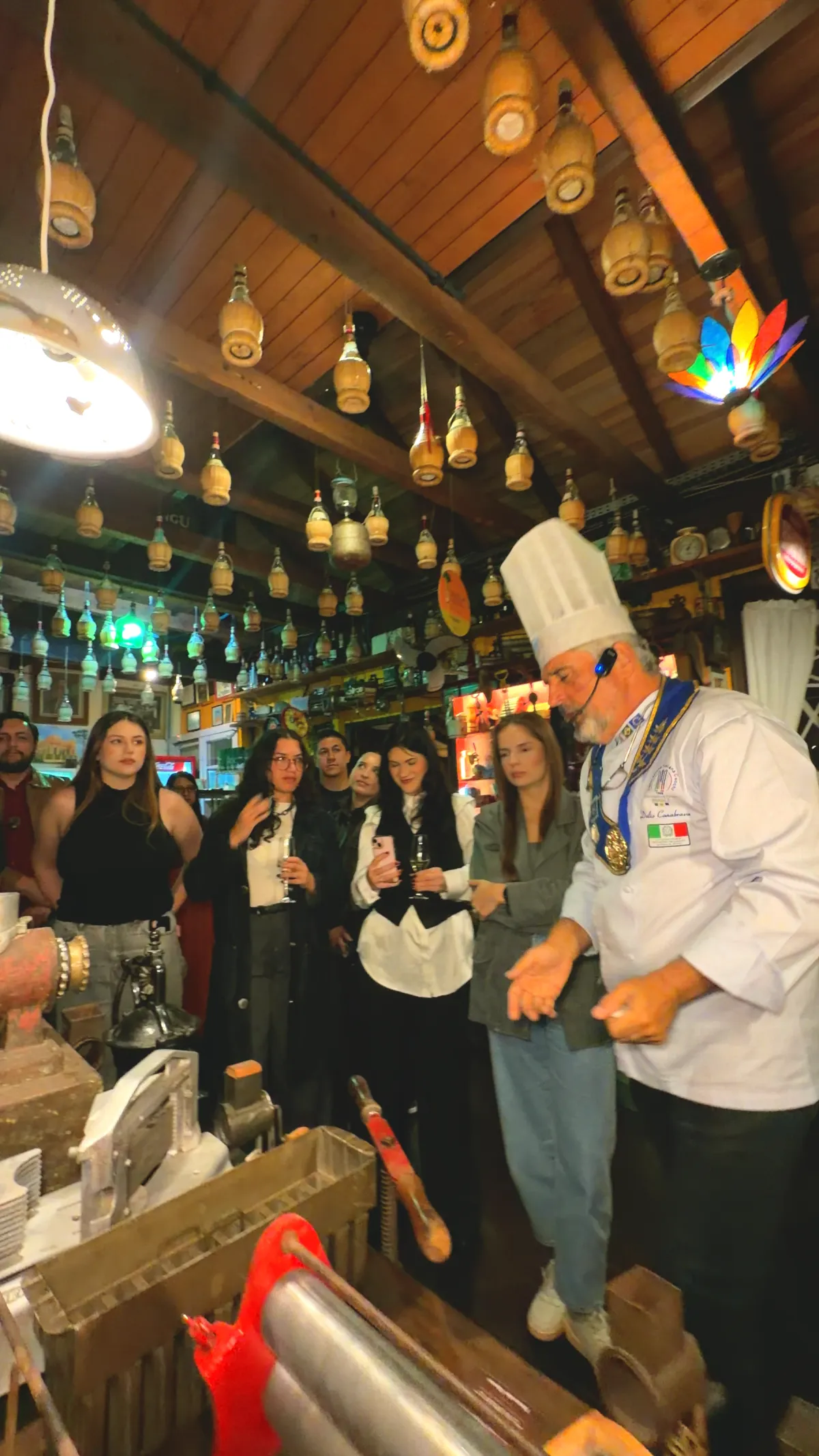 Délio no deck rodeado de convidados, sob o teto de garrafas de chianti