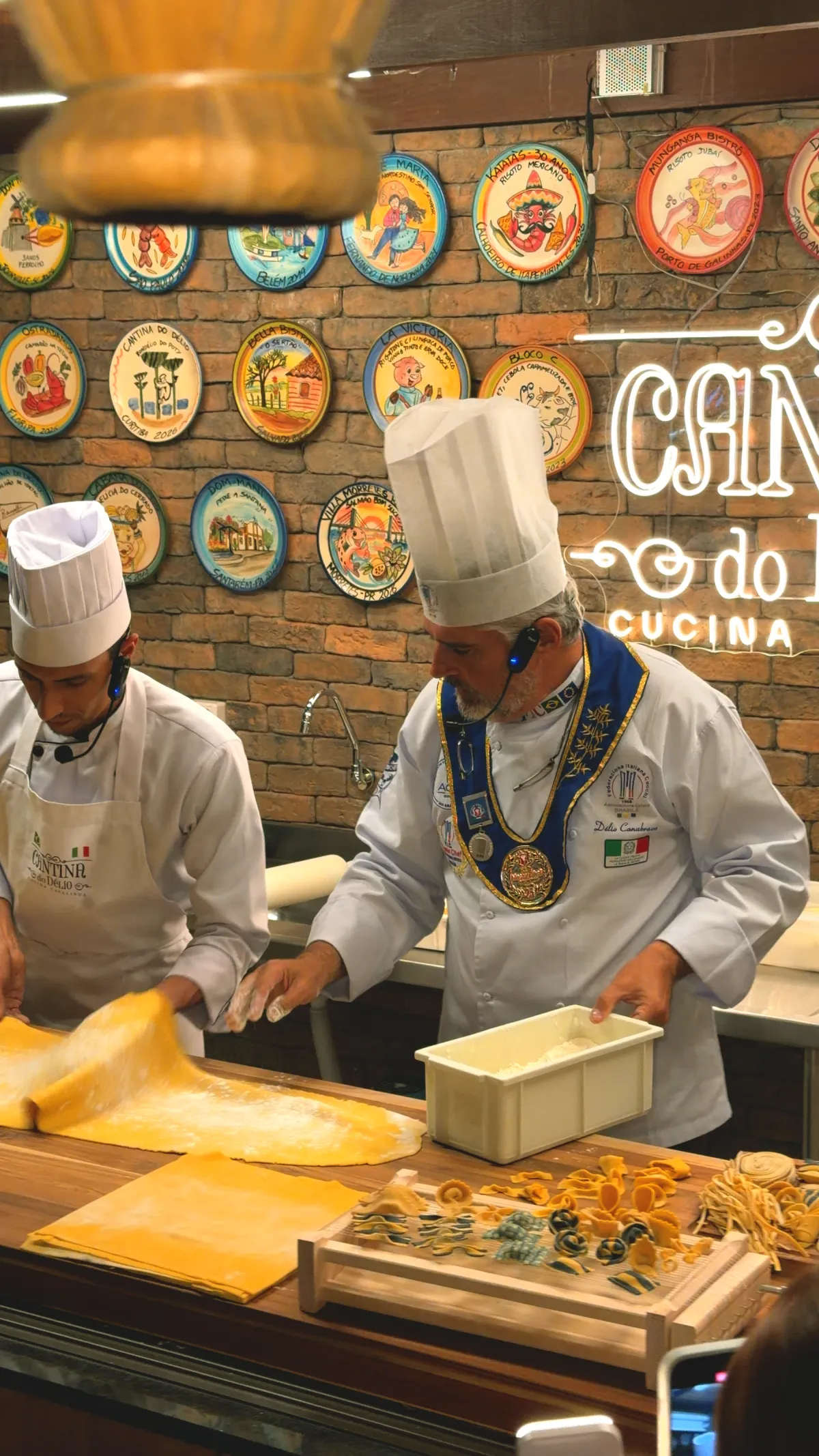 Délio e cozinheiros na bancada, com a parede de pratos e o letreiro da Cantina