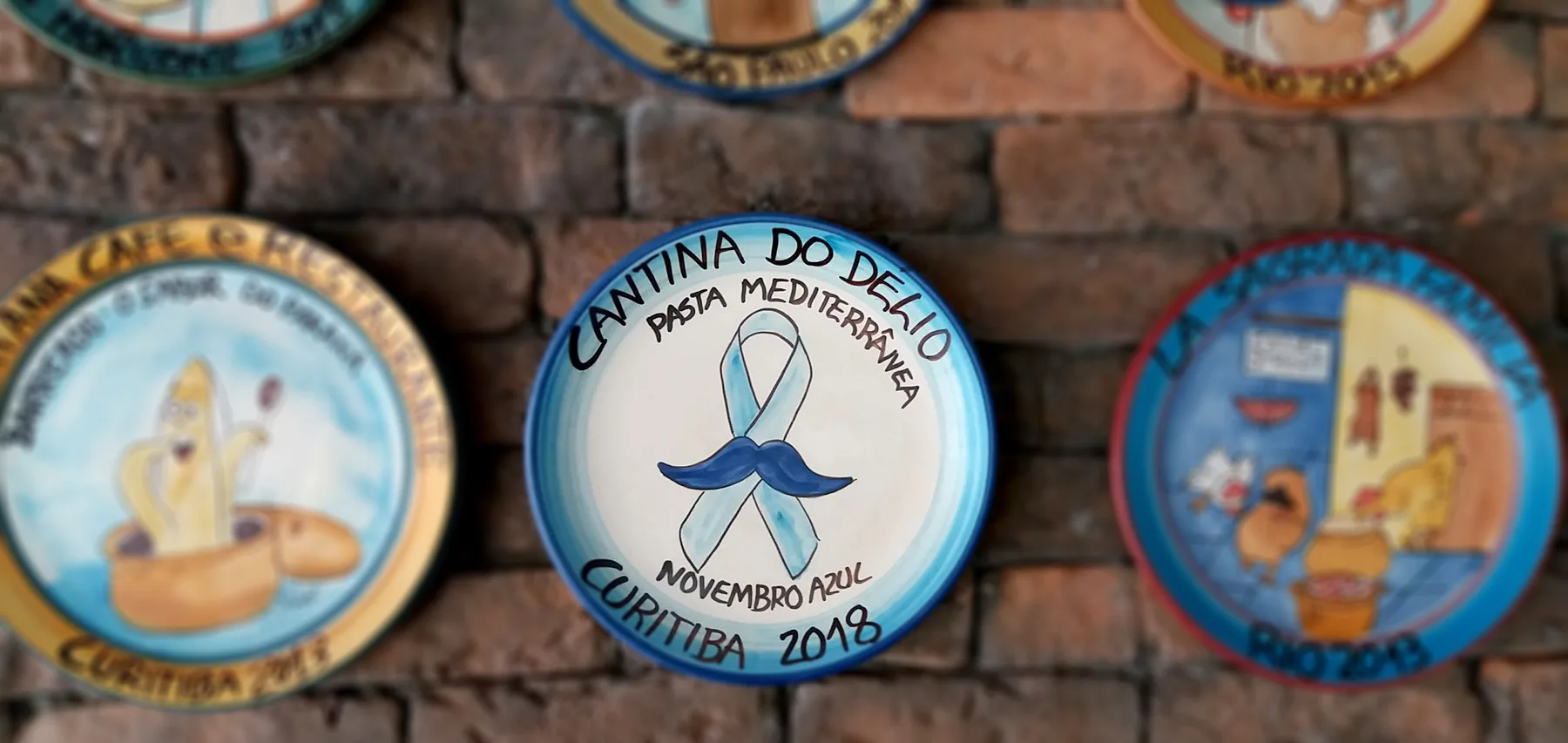Cerâmicas da Boa Lembrança da Cantina penduradas na parede de tijolos, entre elas a do Novembro Azul de 2018